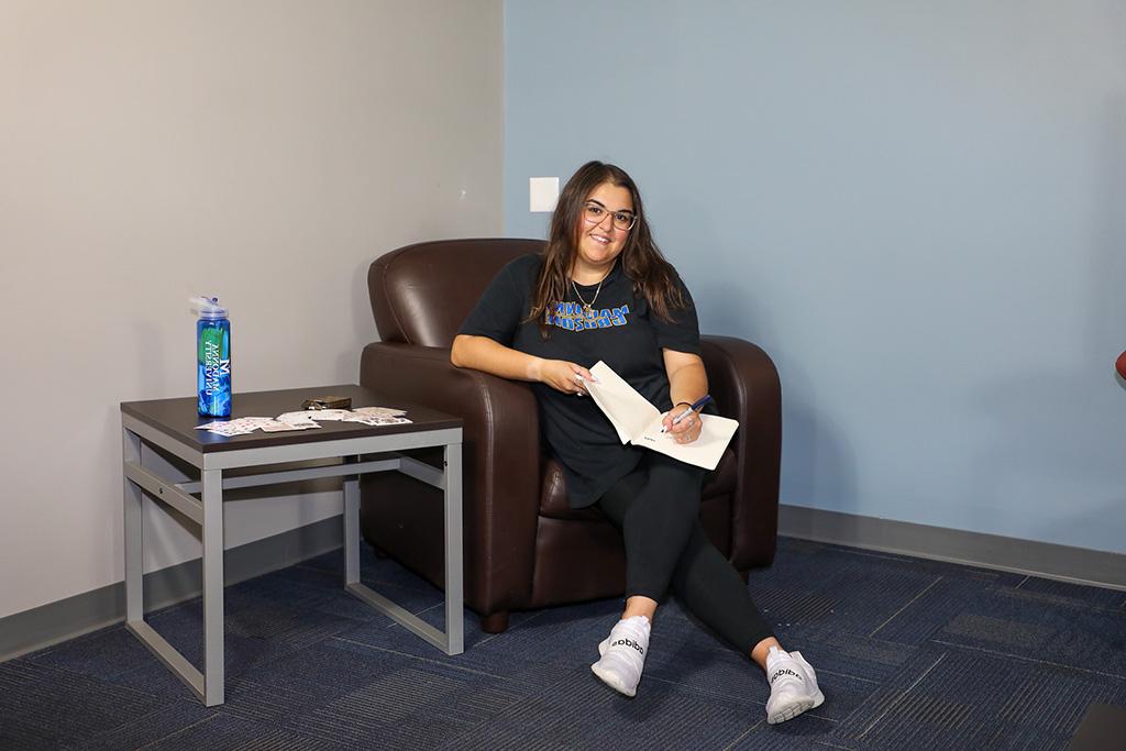 a female sitting in a chair in the residence halls doing homework smiles for the camera